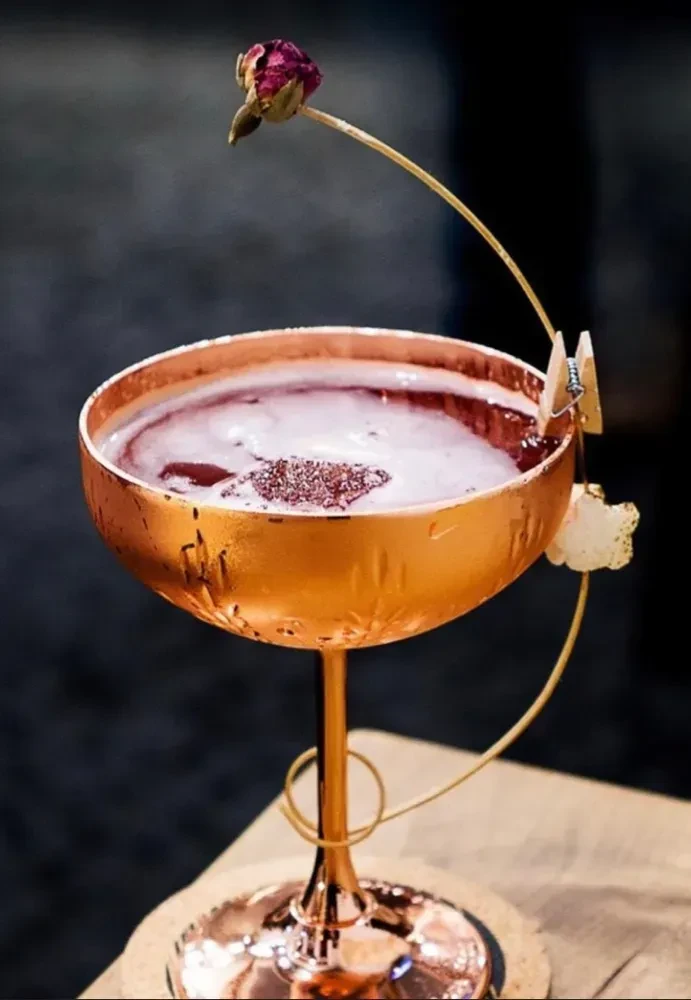 A copper coupe glass with a drink and a dried rose garnish.