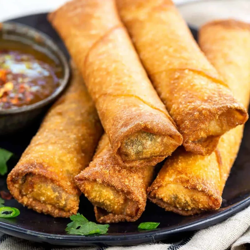 Golden-brown crispy egg rolls on a black plate with dipping sauce.