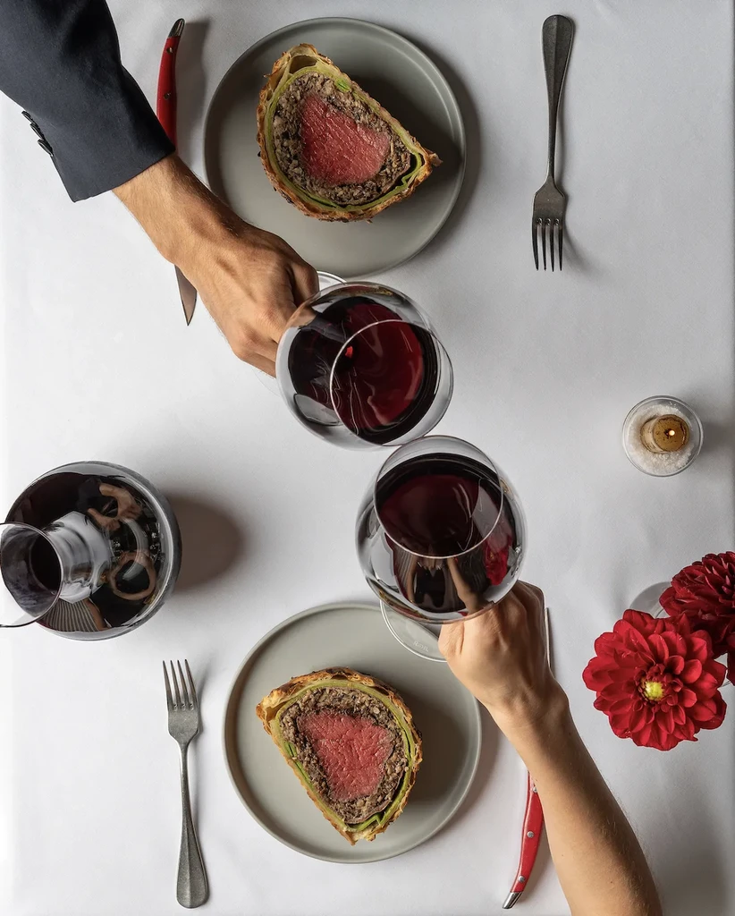 Two hands clinking red wine glasses over Beef Wellington.