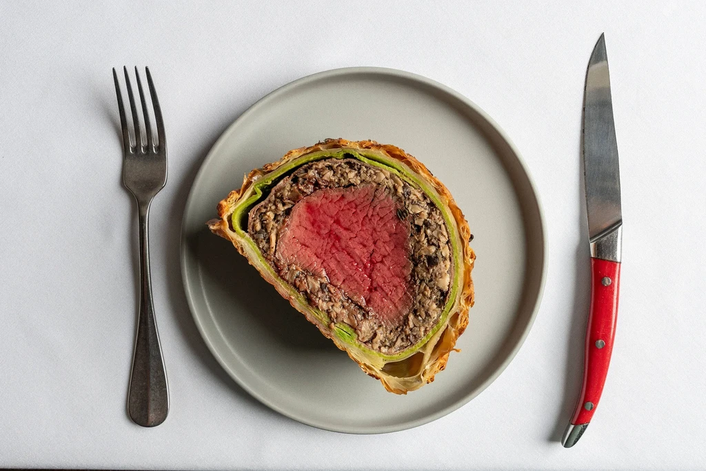 Beef Wellington slice with fork and knife on white table.