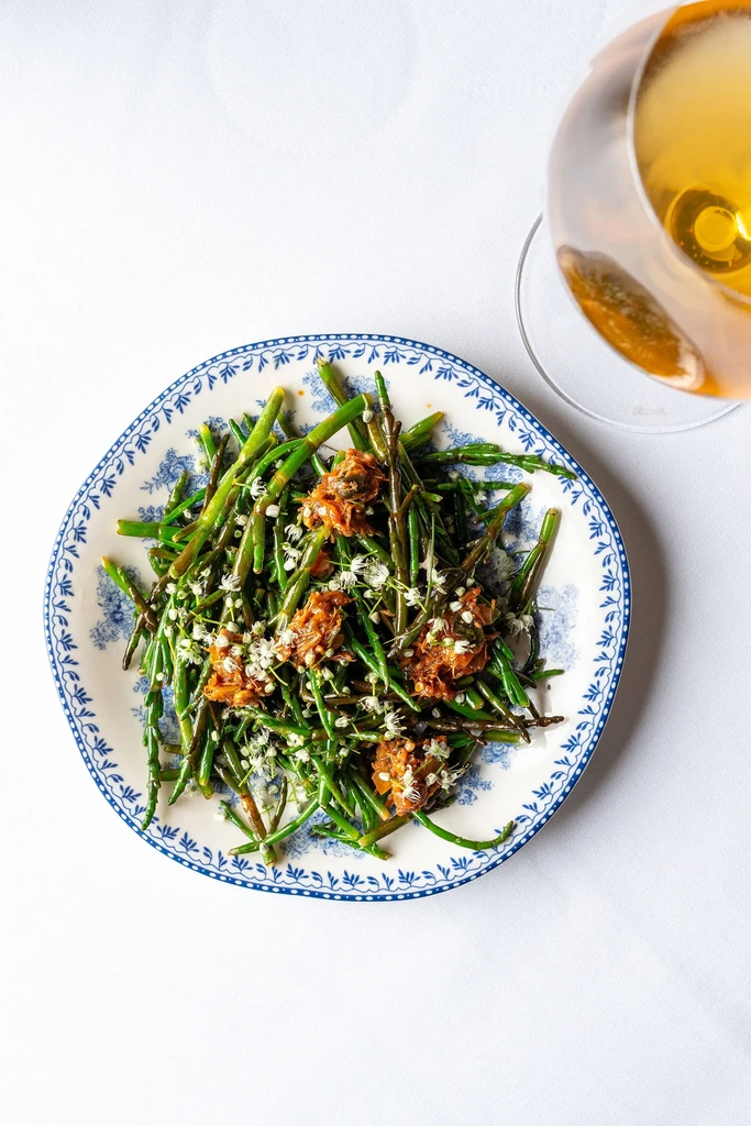 A plate of green vegetables with brown topping and white flowers, next to wine.