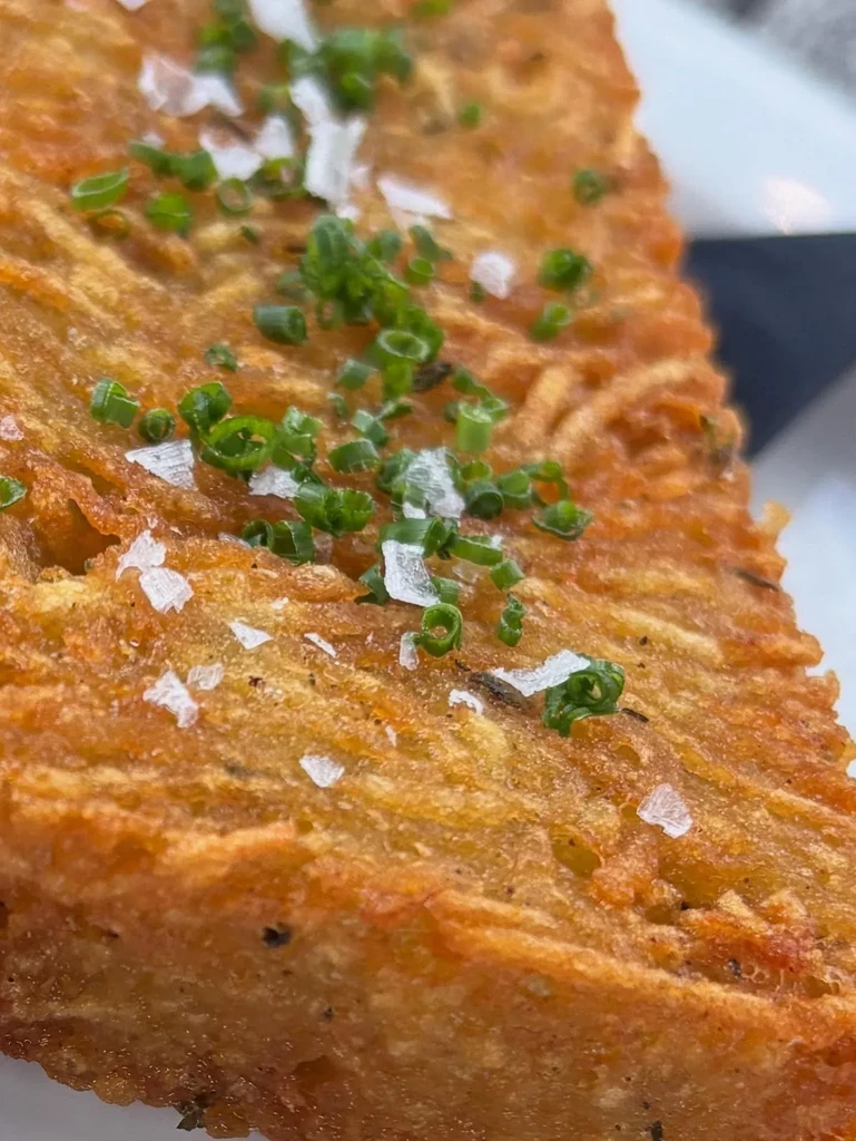 A close-up of a crispy hash brown topped with chives and salt flakes.