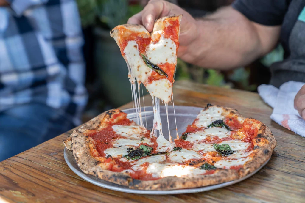 Hand lifts cheesy slice from Margherita pizza on a wooden table.