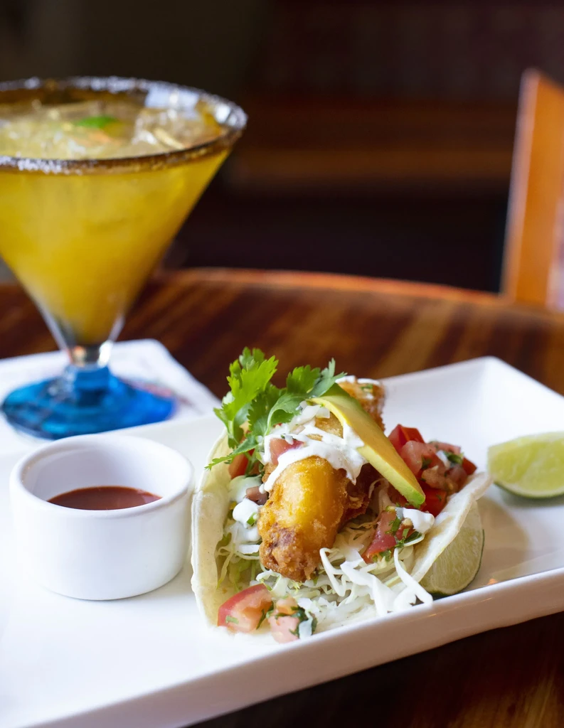 Fish taco with avocado, shredded cabbage, salsa, and a drink.