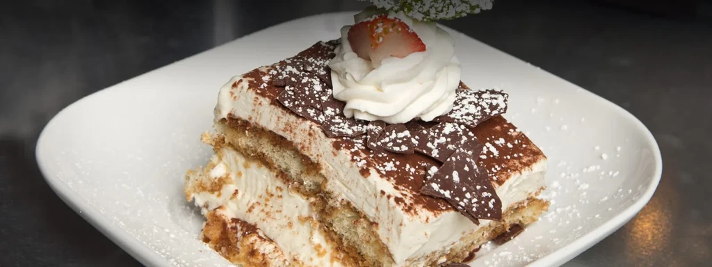 A slice of tiramisu with whipped cream, strawberry, and chocolate shavings.