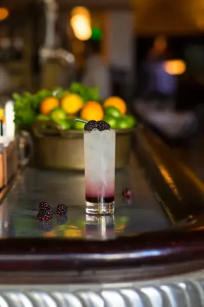 A blackberry cocktail on a bar, with fresh fruit in background.