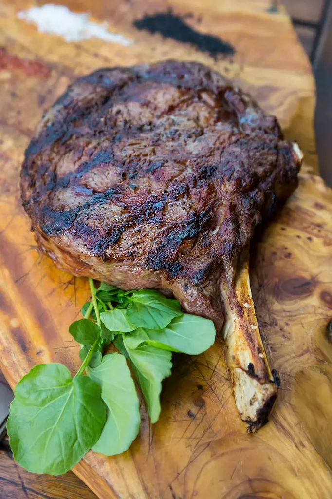 A grilled tomahawk steak with greens on a wooden cutting board.