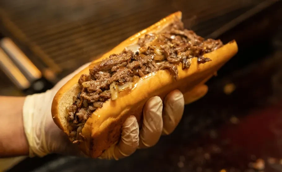 A gloved hand holds a cheesesteak sandwich filled with meat and onions.