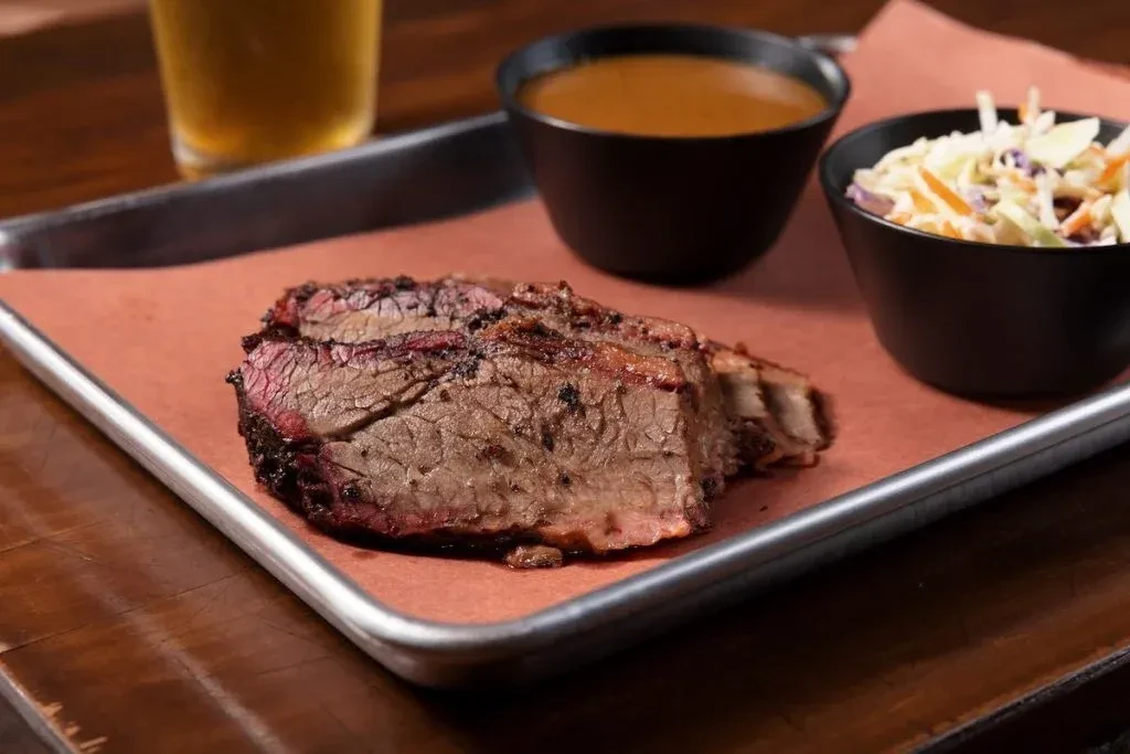 Sliced brisket with sauce and coleslaw on a metal tray.