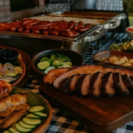 A buffet table with sliced meat, sausages, and side dishes on a checkered tablecloth.