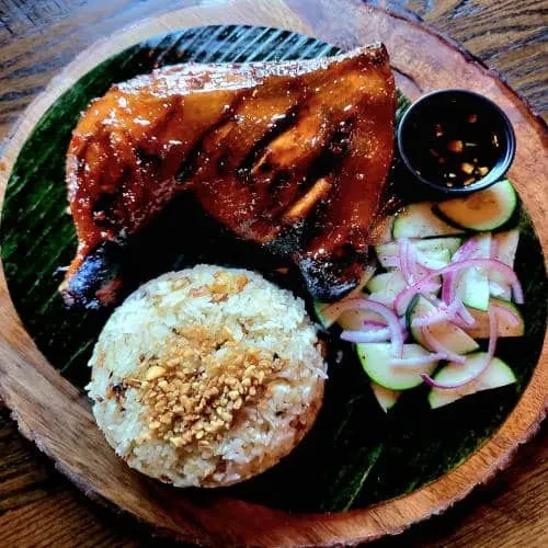 Grilled chicken, rice, cucumber salad, and dipping sauce on a wooden plate.