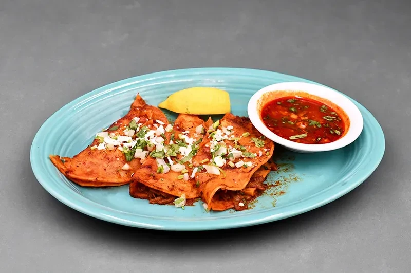Three red birria tacos with a side of sauce and a lemon wedge.