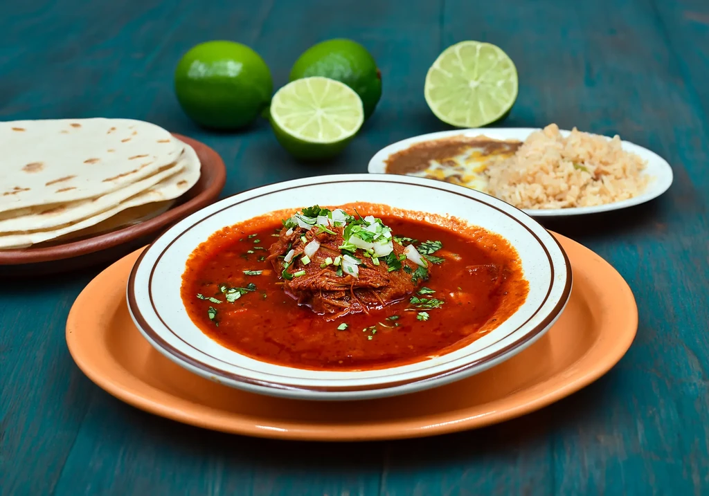 Birria soup with tortillas, rice, beans, and limes on a blue table.