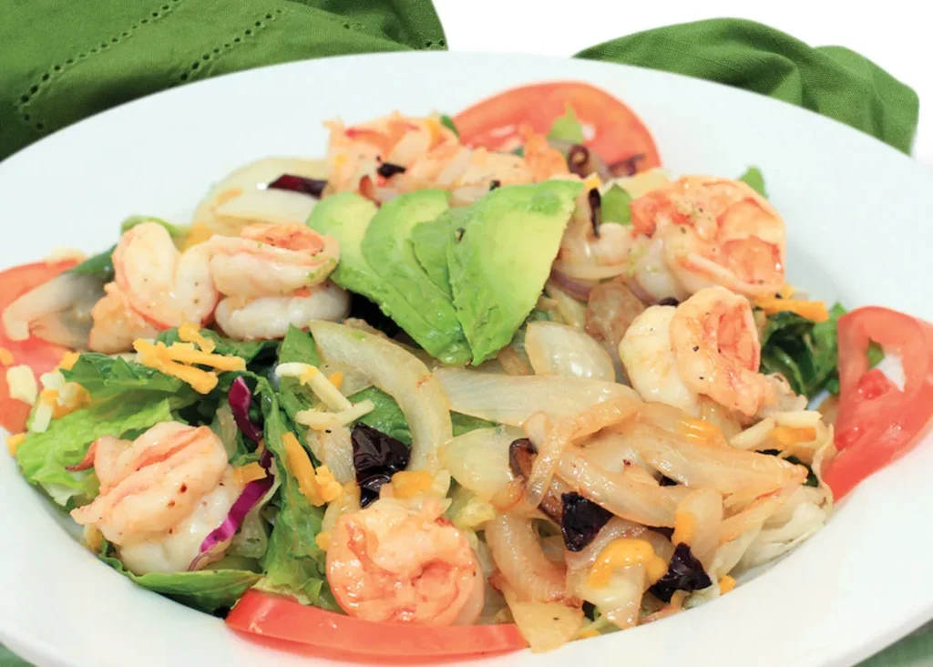 A white bowl filled with shrimp, avocado, lettuce, tomatoes, and onions.