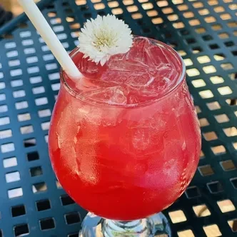 Red drink in stemmed glass with ice, straw, white flower garnish.