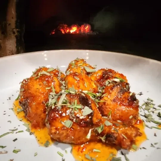 Glazed chicken with herbs in a bowl, wood-fired oven in background.