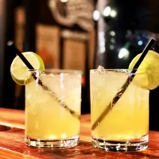 Two cocktails with lime, ice, and straws on a wooden bar.