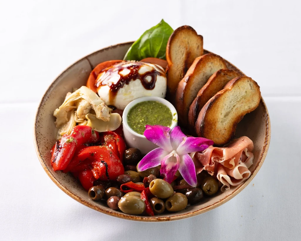 Rustic platter with mozzarella, prosciutto, roasted vegetables, olives, bread, and orchid.