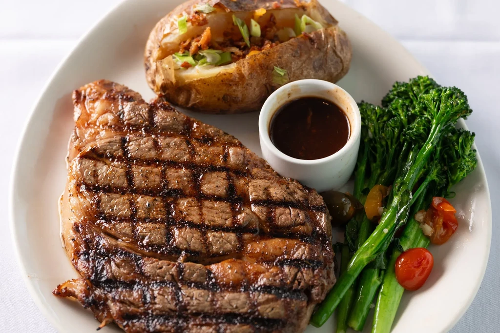 Grilled steak, loaded baked potato, broccolini, and brown sauce on oval plate.