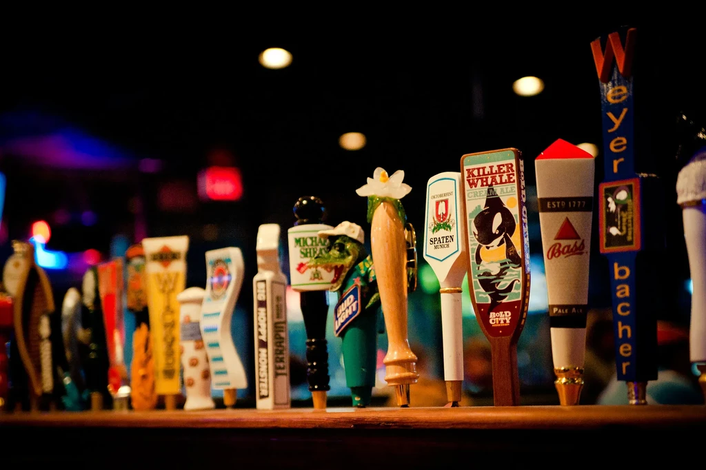 Colorful beer tap handles on a wooden bar against blurred lights.