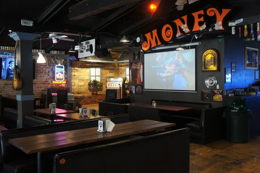 Eclectic bar interior with booths, neon signs, jukebox, and projection screen.