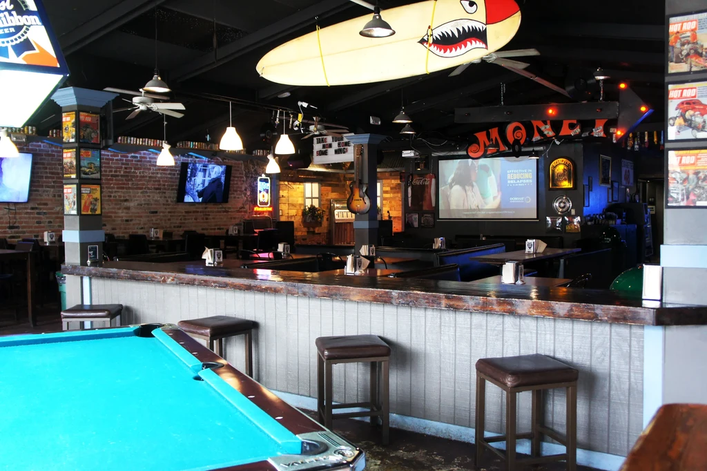 Industrial-rustic bar interior with pool table, prominent surfboard, and projector screen.