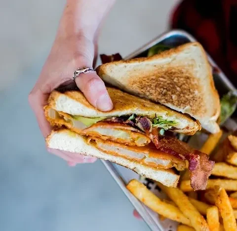 Hand holds a cut-in-half sandwich with fried patty, bacon, and fries.