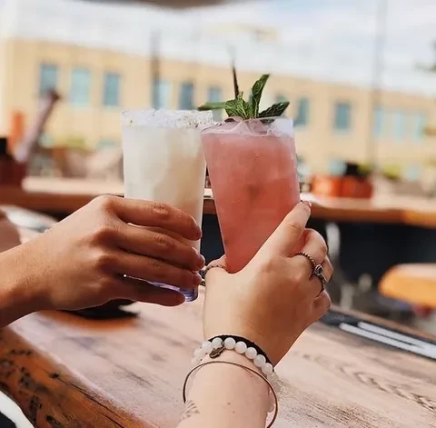 Two hands clinking colorful white and pink cocktails outdoors.