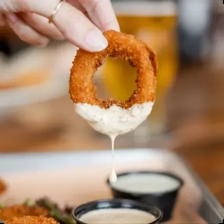 Hand holds crispy onion ring dripping creamy white sauce.