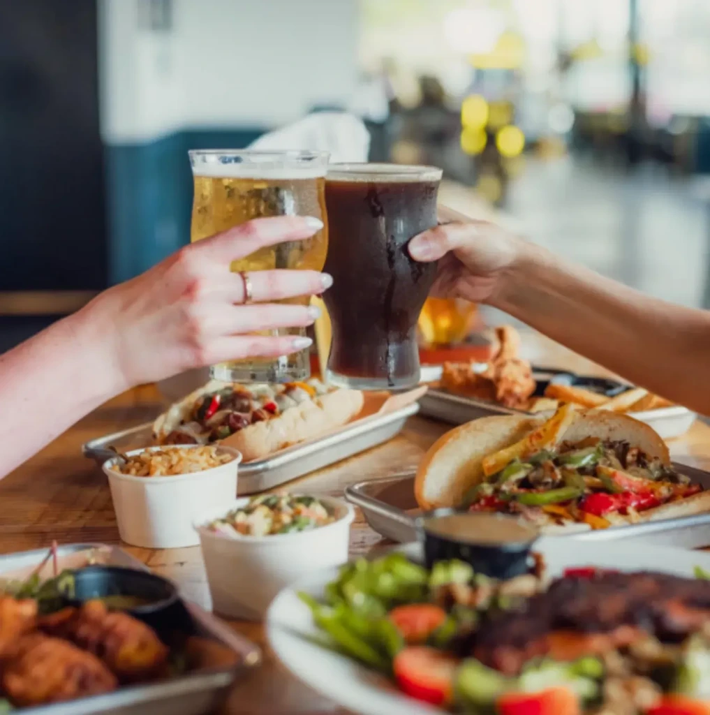 Hands clinking light and dark beers over a restaurant table of food.