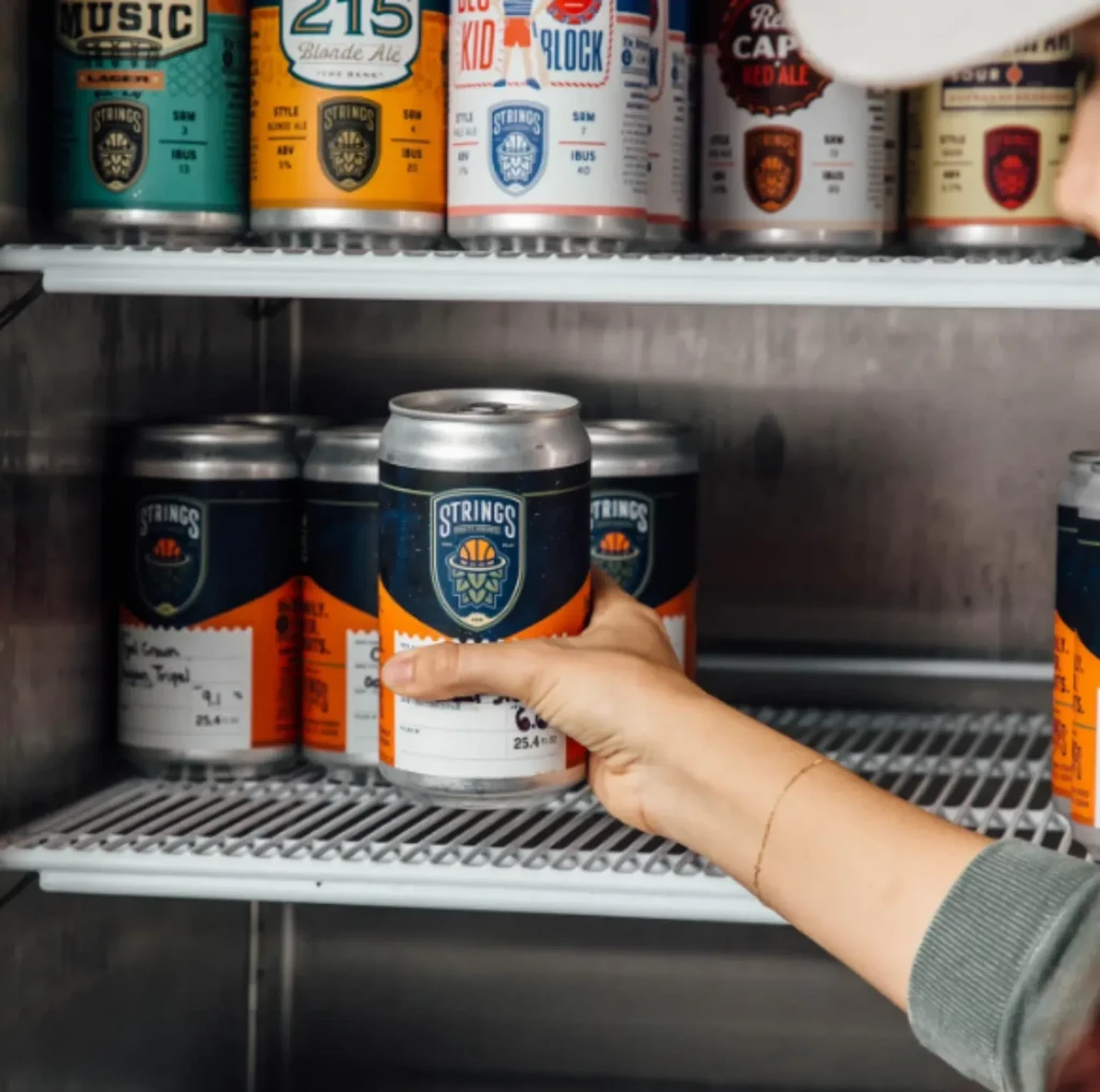 Hand reaching for a 'STRINGS' craft beer can in a cooler.