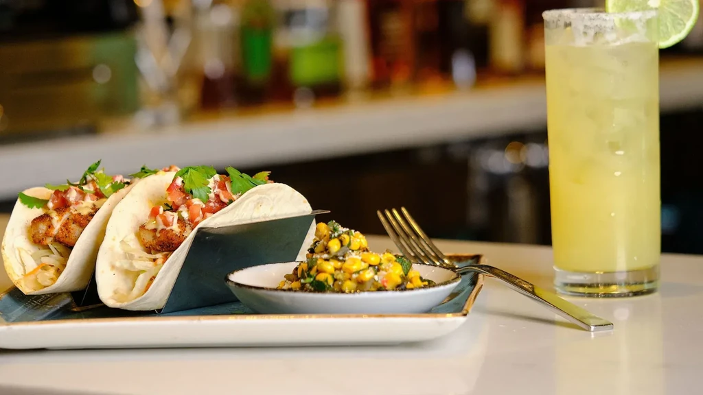 Two fish tacos, corn salad, and a margarita on a white counter.