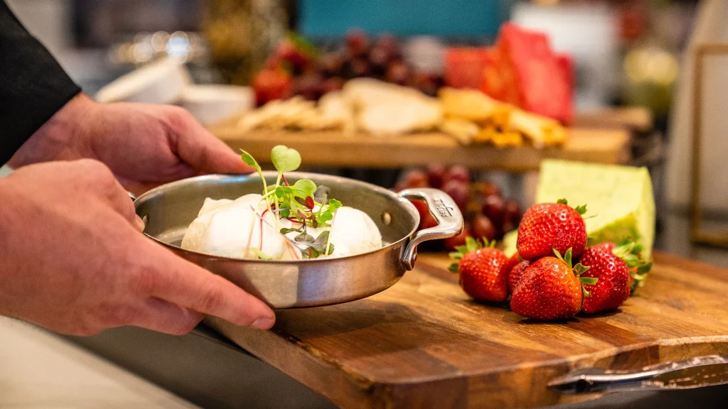 Hands present mozzarella, microgreens, strawberries, and a cheese buffet.