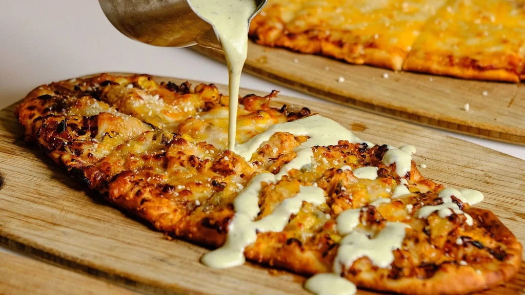 Oval flatbread pizza with cheese, onions, and green sauce pouring.