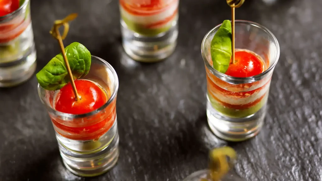 Shot glasses with layered green, white, red appetizers, cherry tomato, basil.
