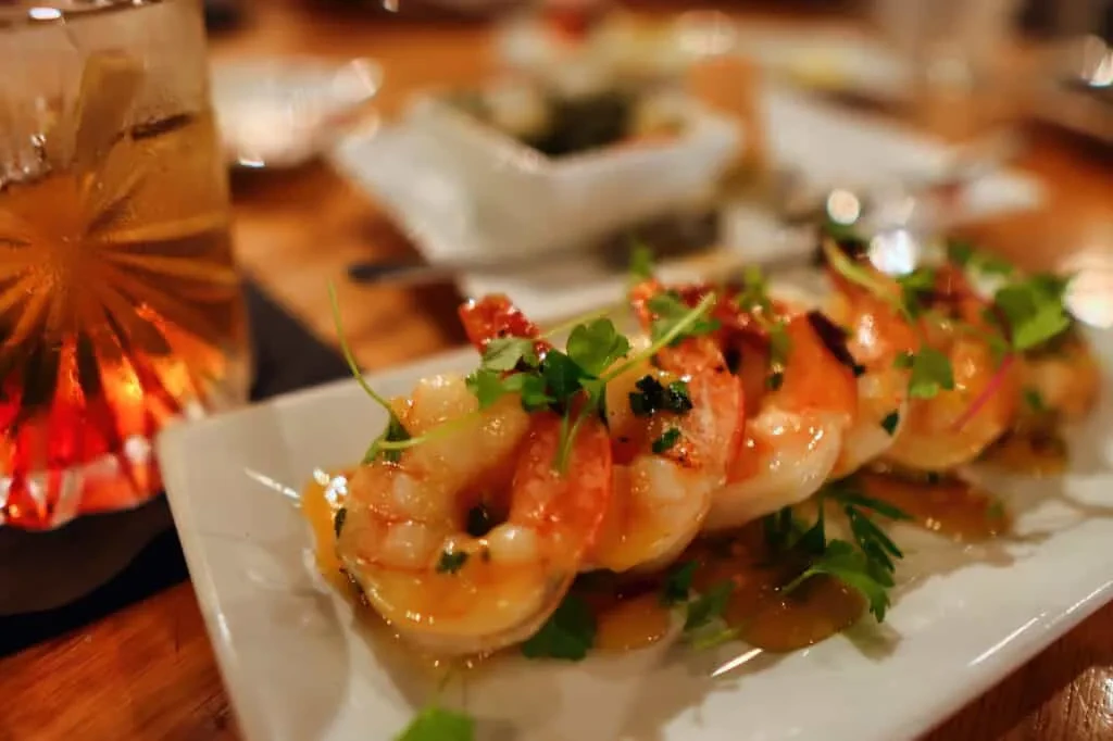 Cooked shrimp dish with herbs and sauce on white plate, cocktail nearby.