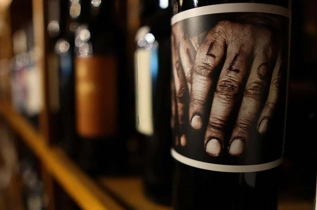 Dark wine bottle label with weathered hands and finger tattoos.