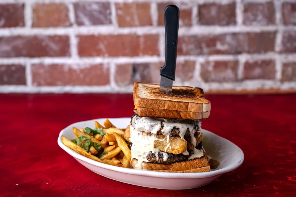 Tall burger between toasted bread with cheese, fried items, knife, and fries.