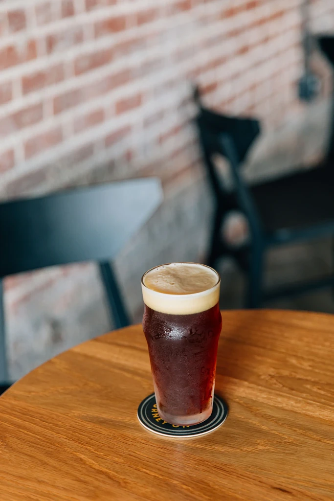 Chilled dark beer with creamy head on wooden table, blurred brick wall.