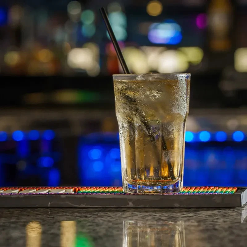 Cold amber drink with ice, straw, on bar mat; blurred background.