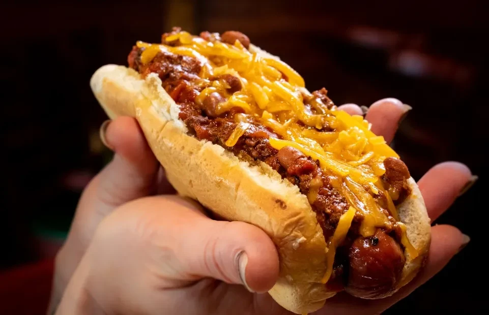 Hand holding a chili cheese hot dog topped with melted cheese.