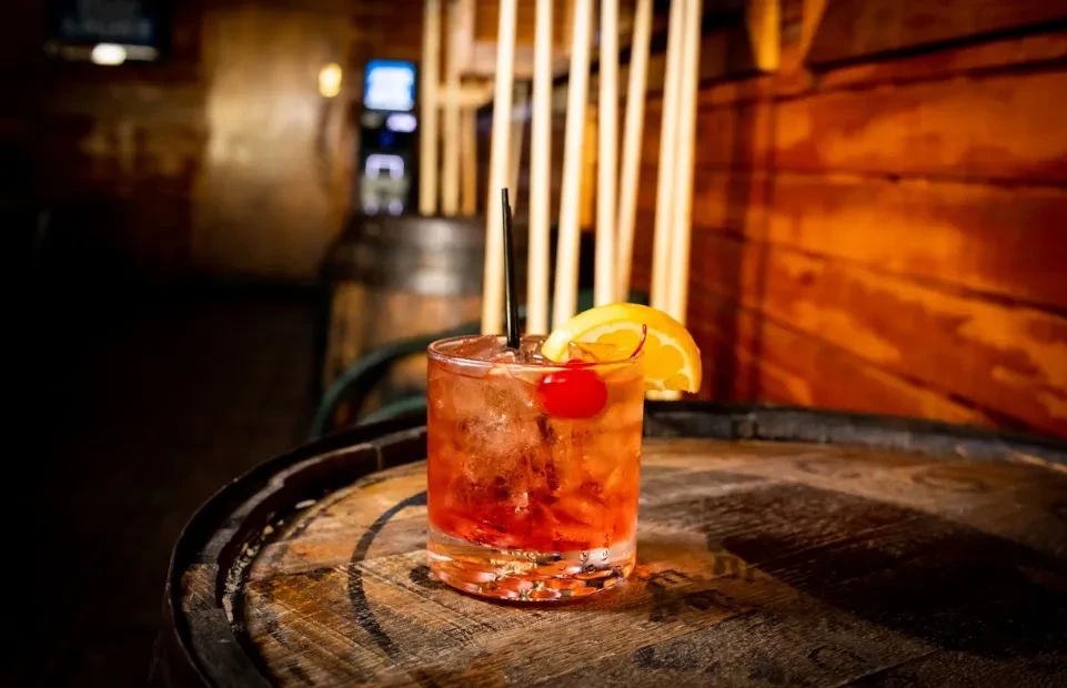 Iced red-orange cocktail with cherry, orange, and straw on a barrel.