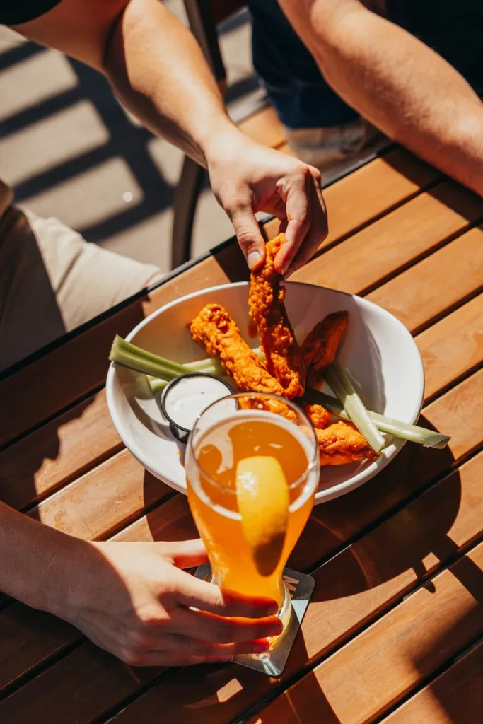 Hands grabbing fried chicken and beer on a sunny wooden table.