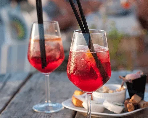 Two large red iced drinks with appetizers on a rustic wooden table.