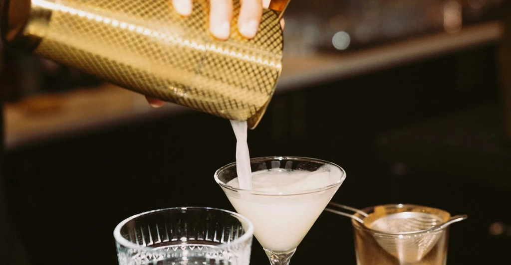 A hand pours a cocktail from a gold shaker into a martini glass.