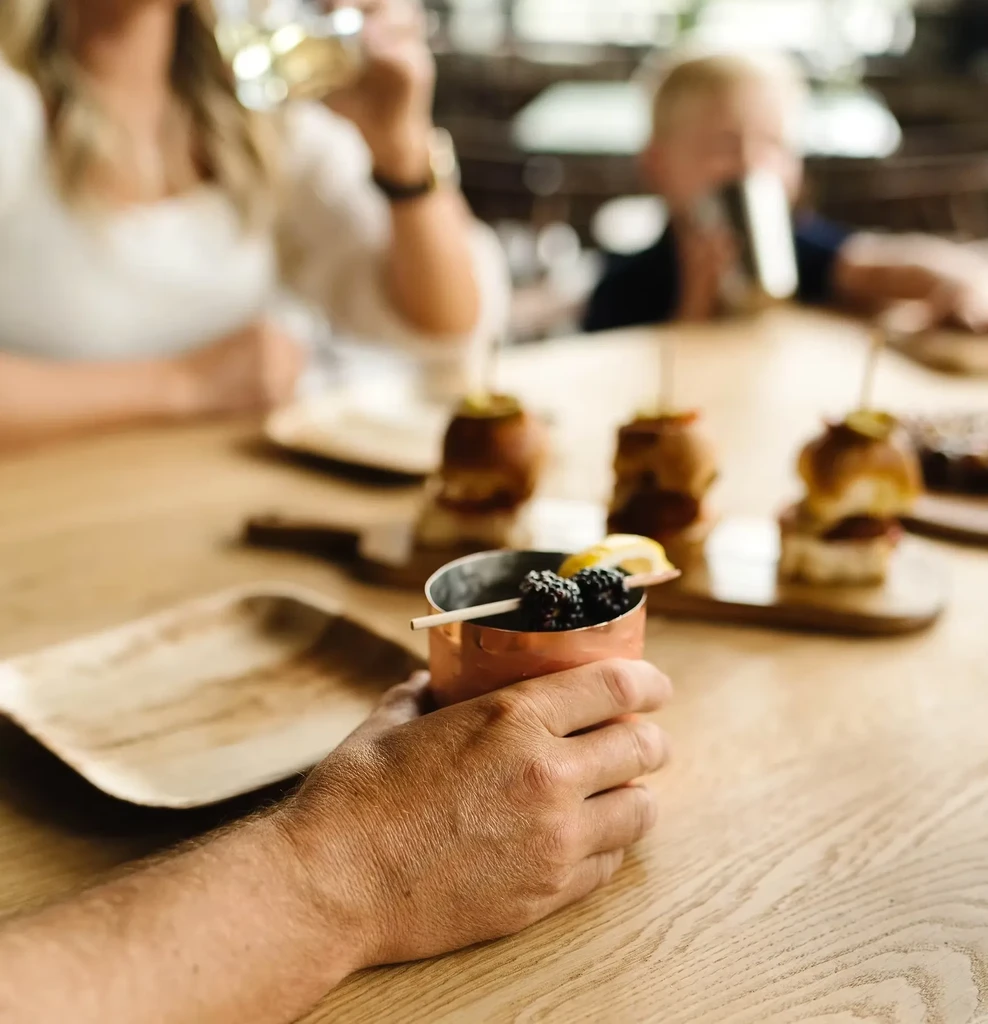 Person holds copper mug cocktail, with sliders and blurred diners behind.