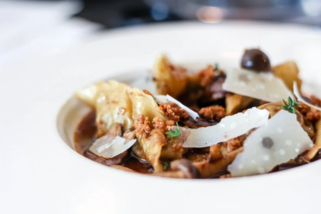 Meticulously plated pasta in dark sauce, garnished with cheese and herbs.