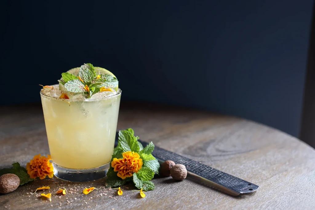 Pale green cocktail, mint, lime, nutmeg, and grater on wooden table.