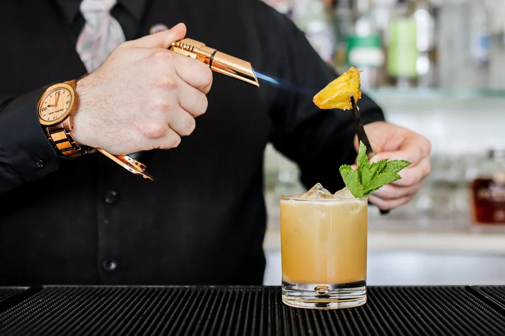 Bartender torches pineapple garnish over a cocktail at a bar.