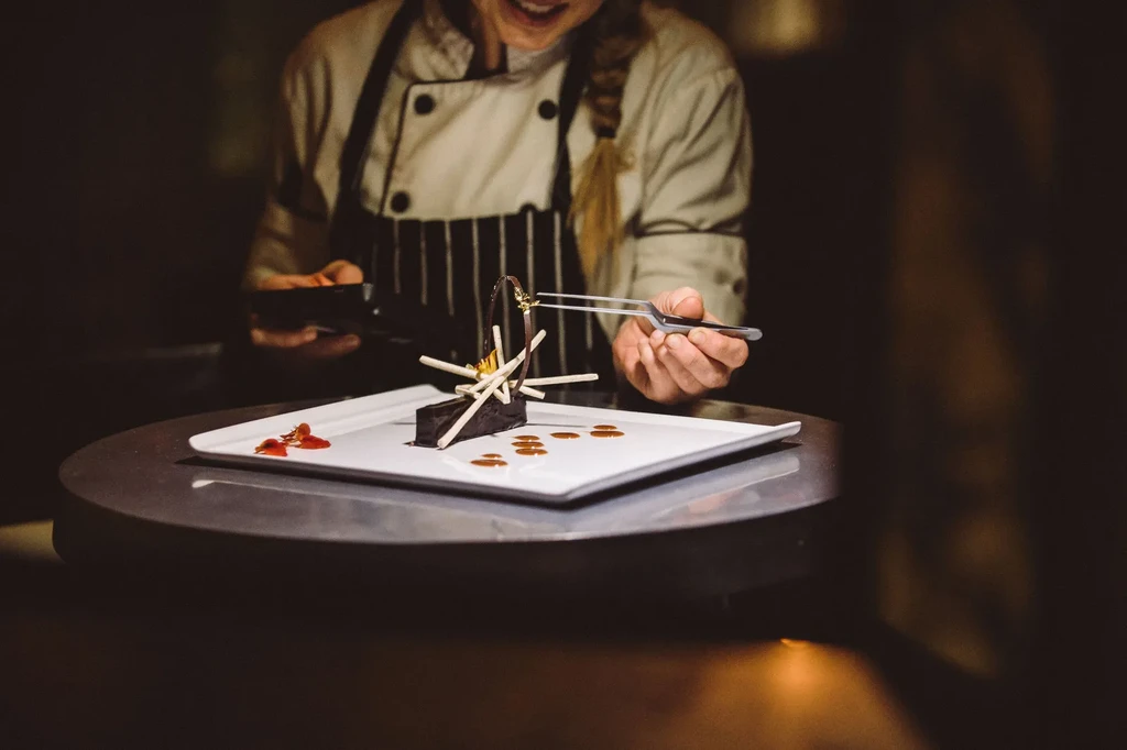 Chef meticulously plates a detailed dark chocolate dessert with tweezers.